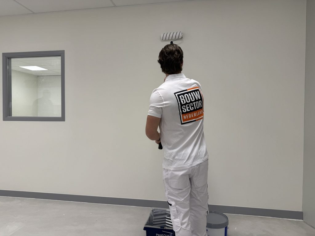 Schilder van Bouwsector Nederland rolt een wand met een verfbak en emmer netjes aan zijn voeten