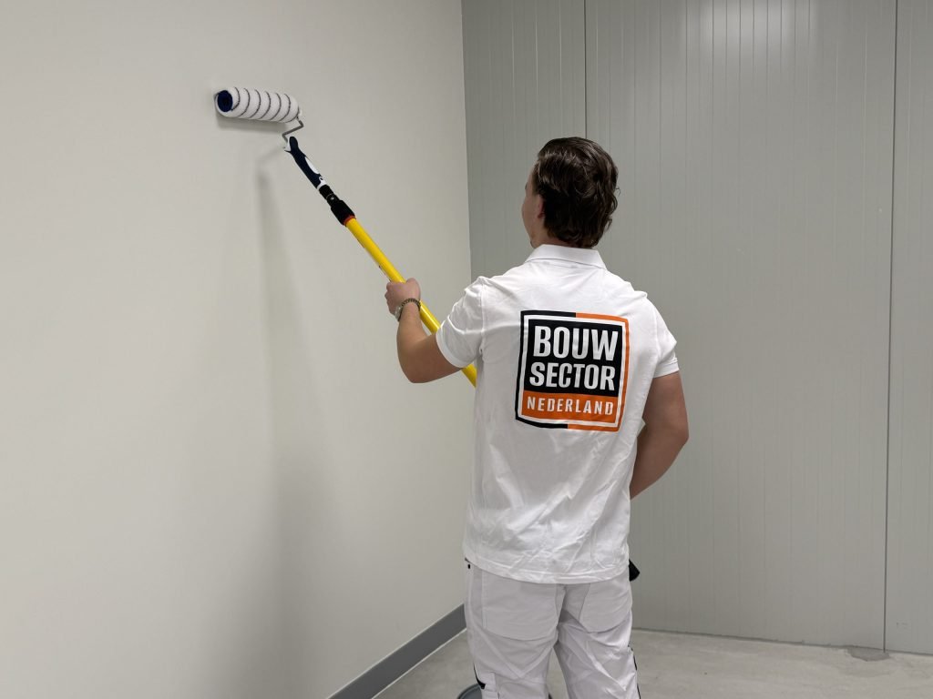Schilder van Bouwsector Nederland zet de roller bovenaan een lichte wand in een lege ruimte