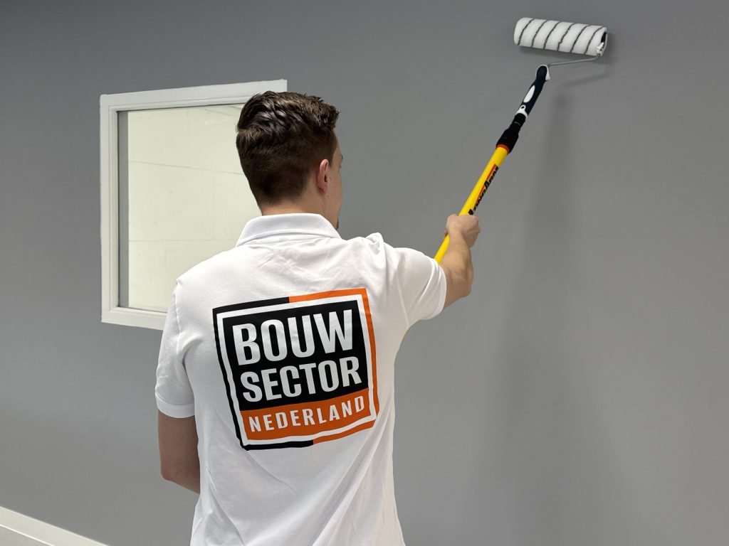 Schilder van Bouwsector Nederland rolt verf op een grijze wand vlak naast een wit raamkozijn